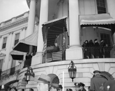 black and white image of FDR standing on a second story balcony giving a speech